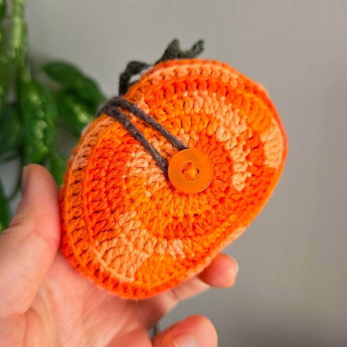 Hand holding a small, round orange crocheted item with a button and string closure, showcasing crocheting skills.