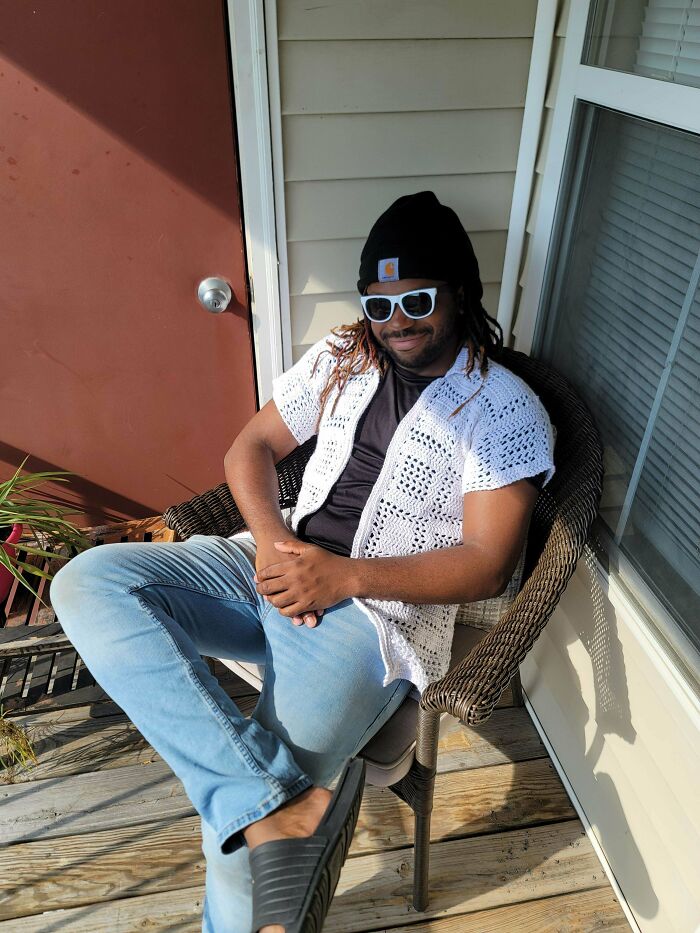 Man wearing white crocheted vest and sunglasses sitting on a chair on a porch showcasing crocheting project outdoors.