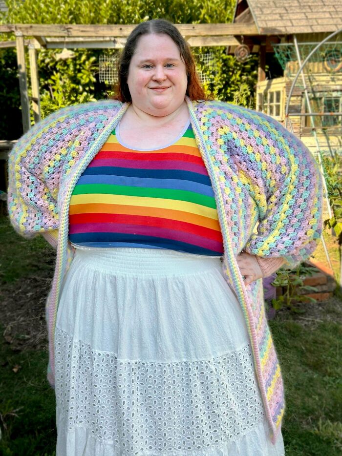 Person wearing a colorful crocheted cardigan and rainbow top, showcasing a handmade crocheting project outdoors in a garden.
