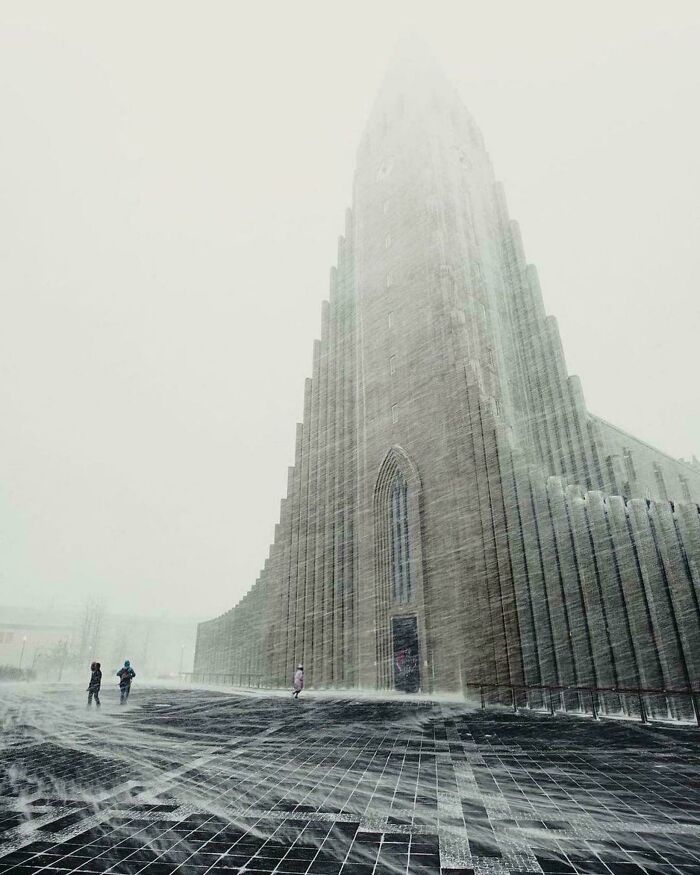 Three people brave the snow in front of an extremely large and stark church, an eerie and majestic structure.