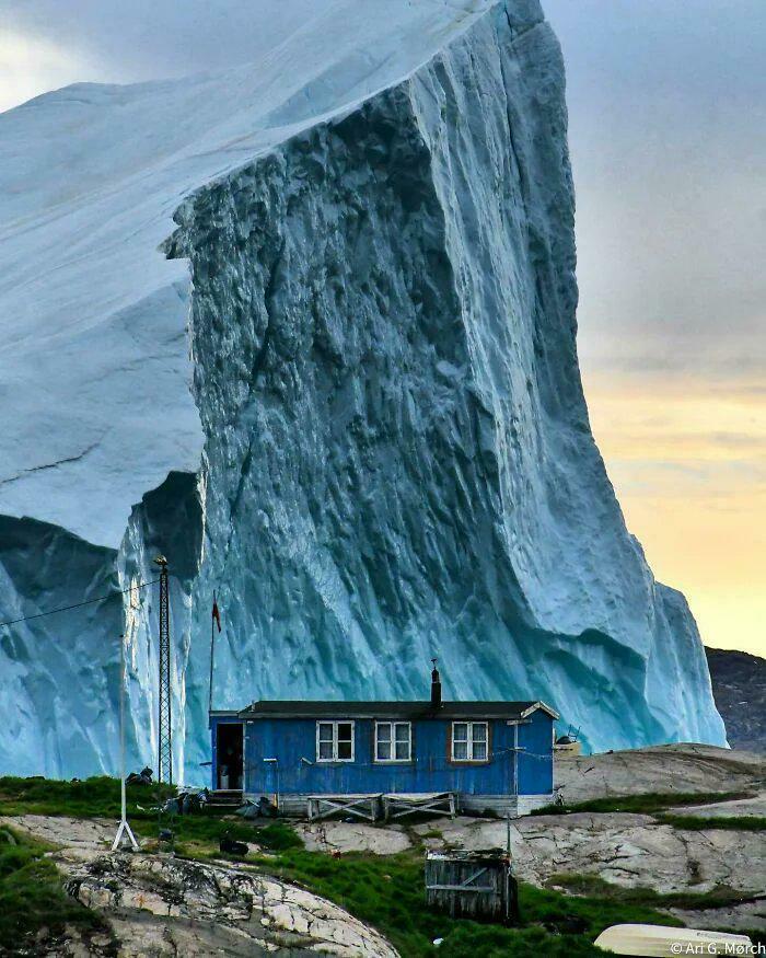An extremely large iceberg towering over a small blue house, highlighting the scary and eerie scale of nature.