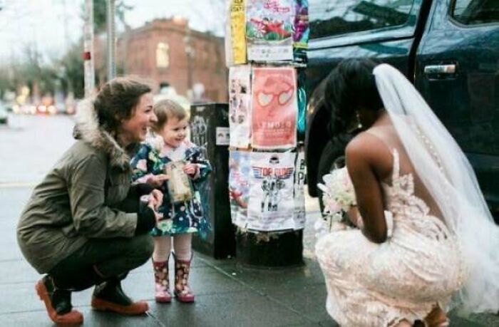 Woman in wedding dress and veil crouching on sidewalk, smiling at happy toddler and mother in heartwarming moment.
