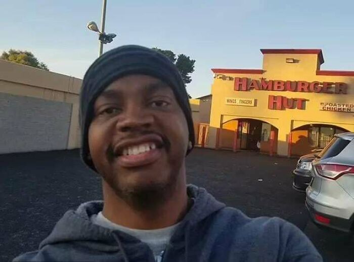 Smiling man wearing a beanie taking a selfie outside Hamburger Hut restaurant in a heartwarming story setting.