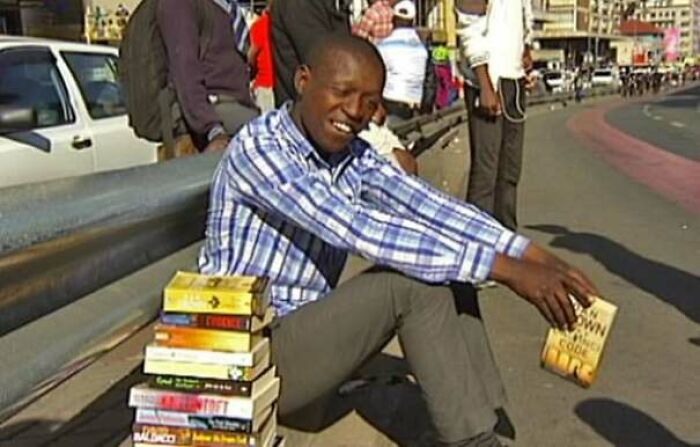 Young man smiling and sitting on sidewalk next to stack of books, spreading heartwarming stories to cleanse negativity.