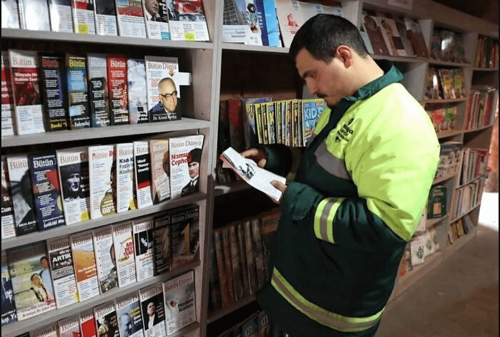 Man in a high-visibility jacket reading a book in a cozy bookstore filled with colorful magazines and stories.