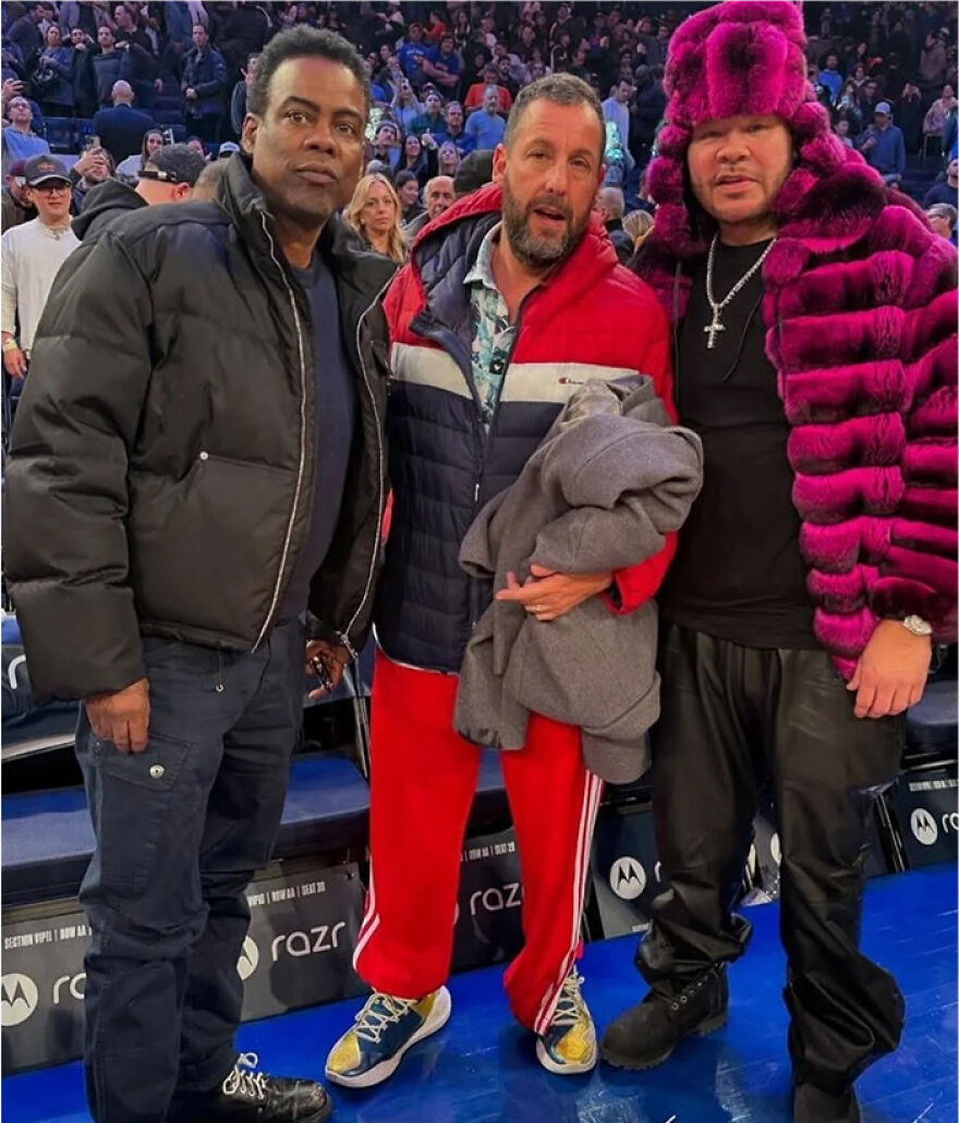 Chris Rock, Adam Sandler, and a man in a pink fur coat posing at an event showcasing Adam Sandler outfits.
