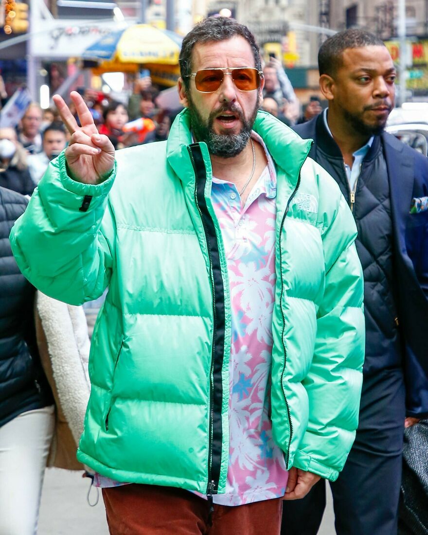 Adam Sandler wearing a mint green puffer jacket and floral shirt, making a peace sign in a busy city street.