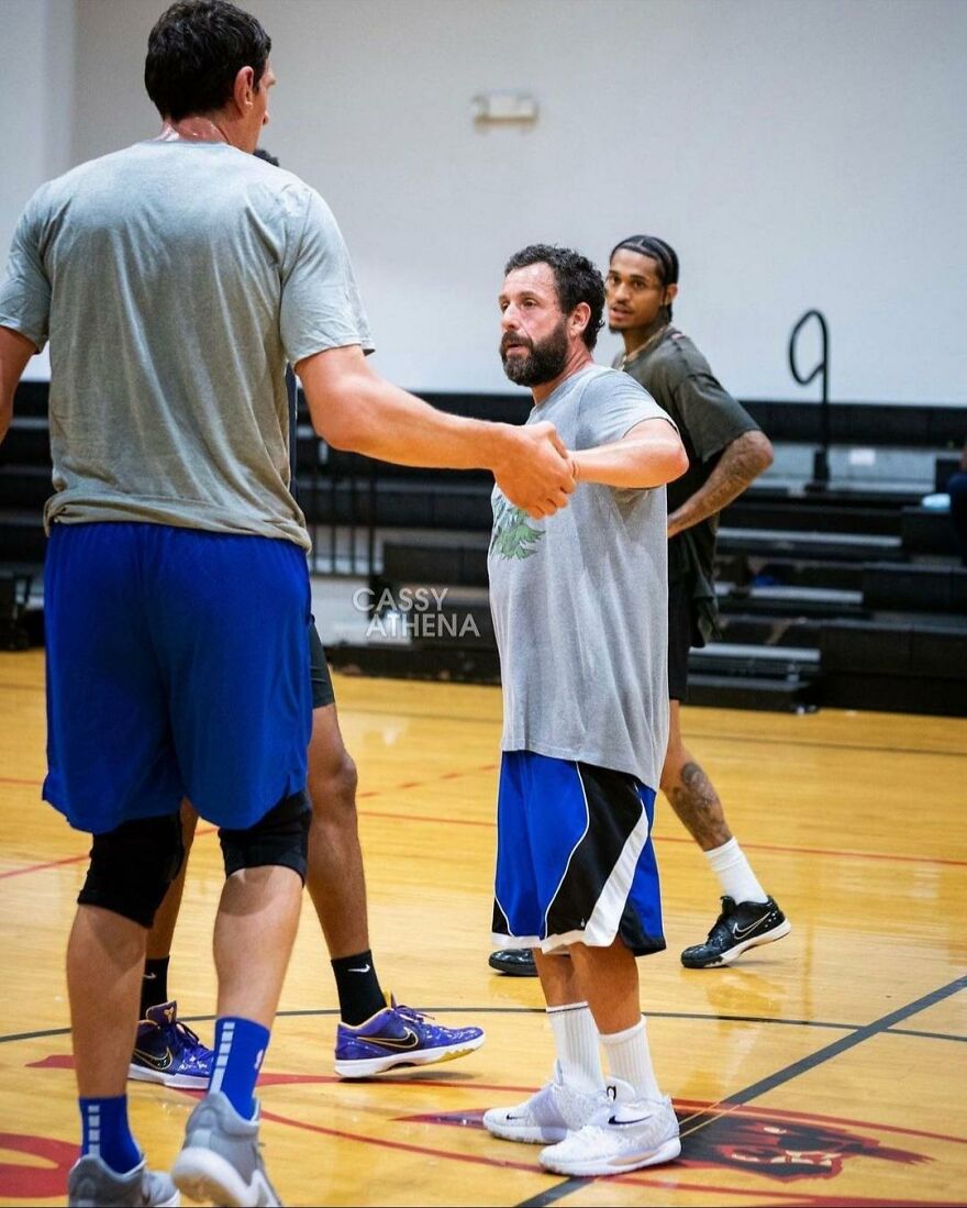 Adam Sandler in casual basketball outfit with gray shirt and blue shorts on an indoor court with teammates.
