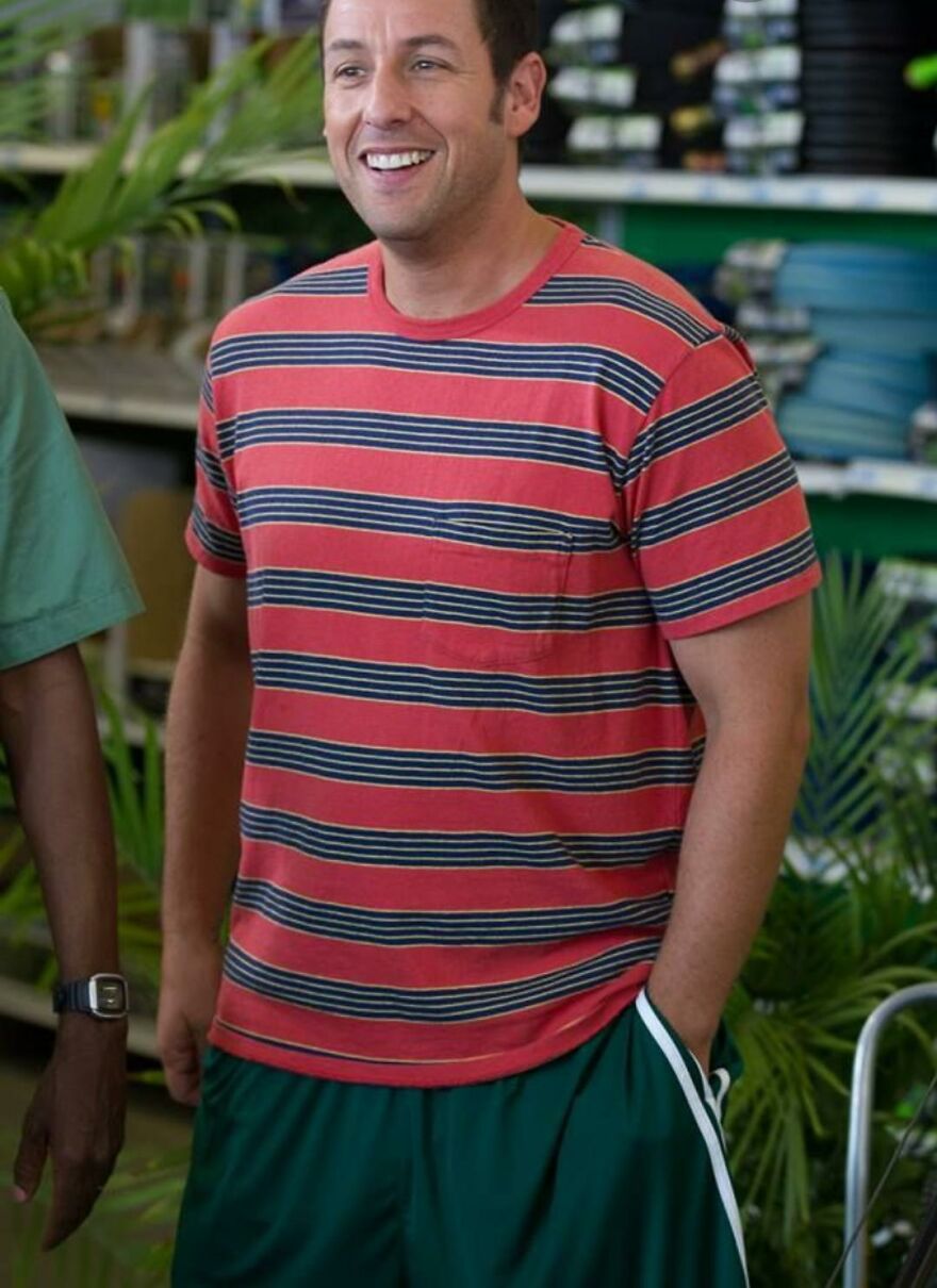 Adam Sandler wearing a red and navy striped t-shirt and green athletic shorts, smiling in a casual setting.