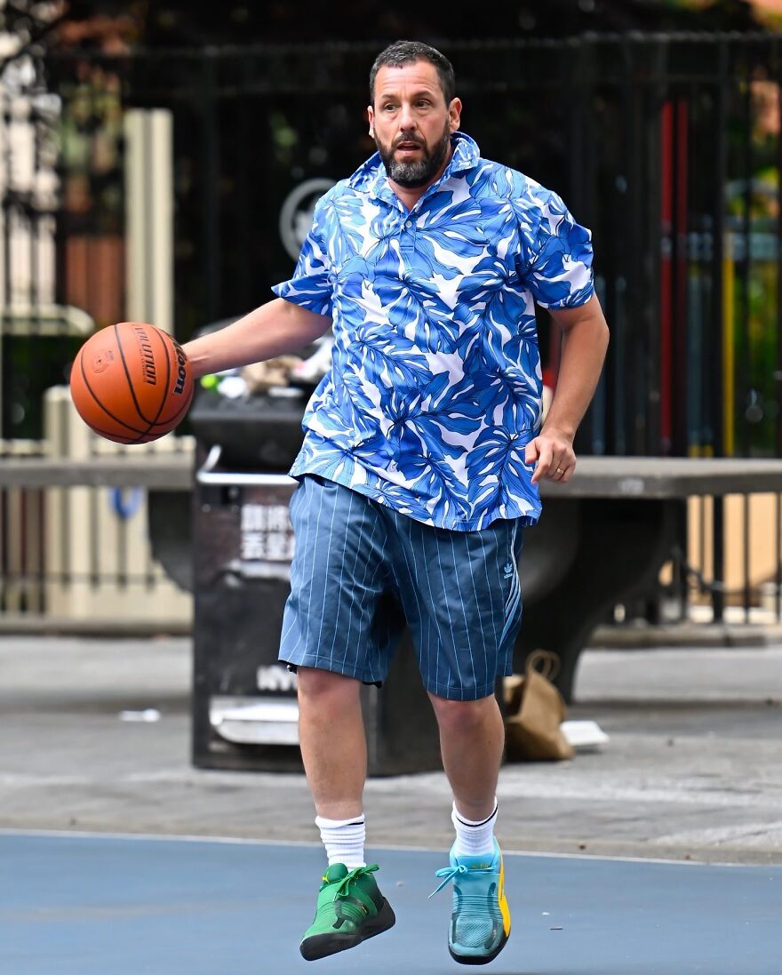Adam Sandler playing basketball outdoors wearing a blue floral shirt and mismatched sneakers, showcasing iconic outfits.