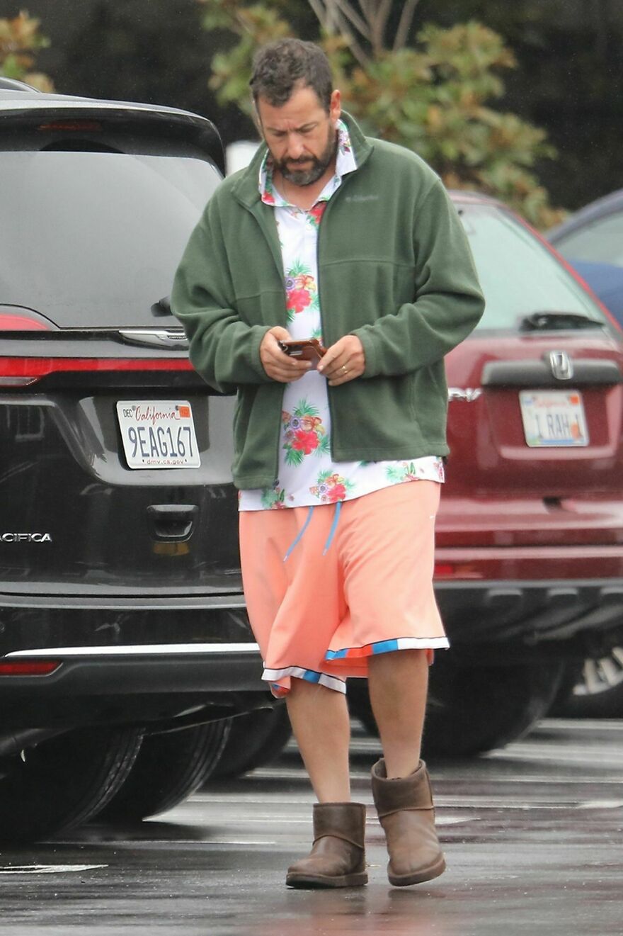 Adam Sandler in colorful floral shirt, green jacket, coral shorts, and brown boots checking phone outdoors.
