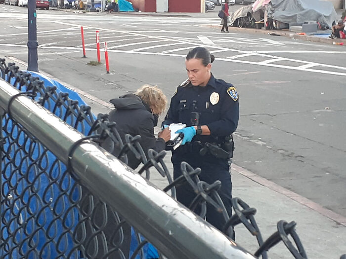 Police officer in uniform assisting a homeless person on a city street reflecting dystopian real-life scenes in the US.