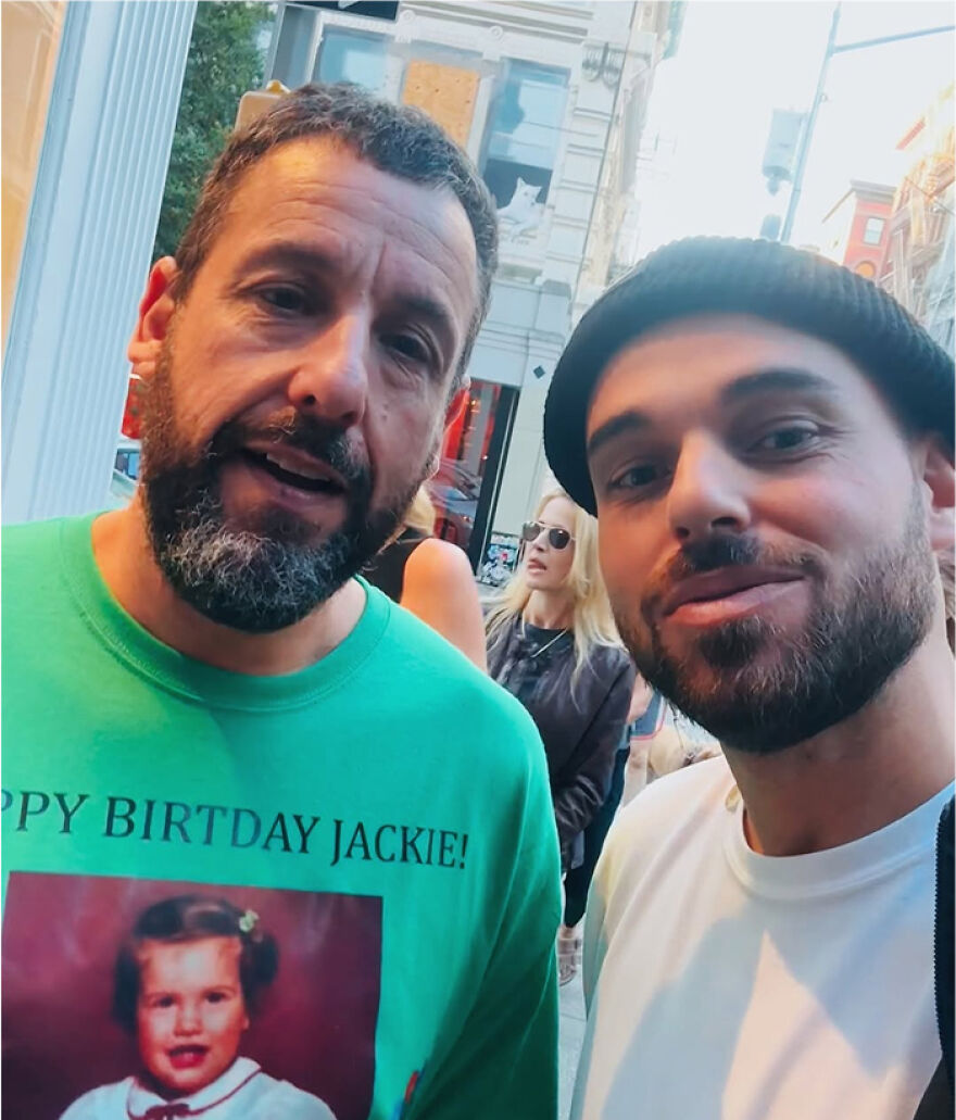 Adam Sandler wearing a green graphic outfit outdoors, posing for a selfie with another man in casual streetwear.