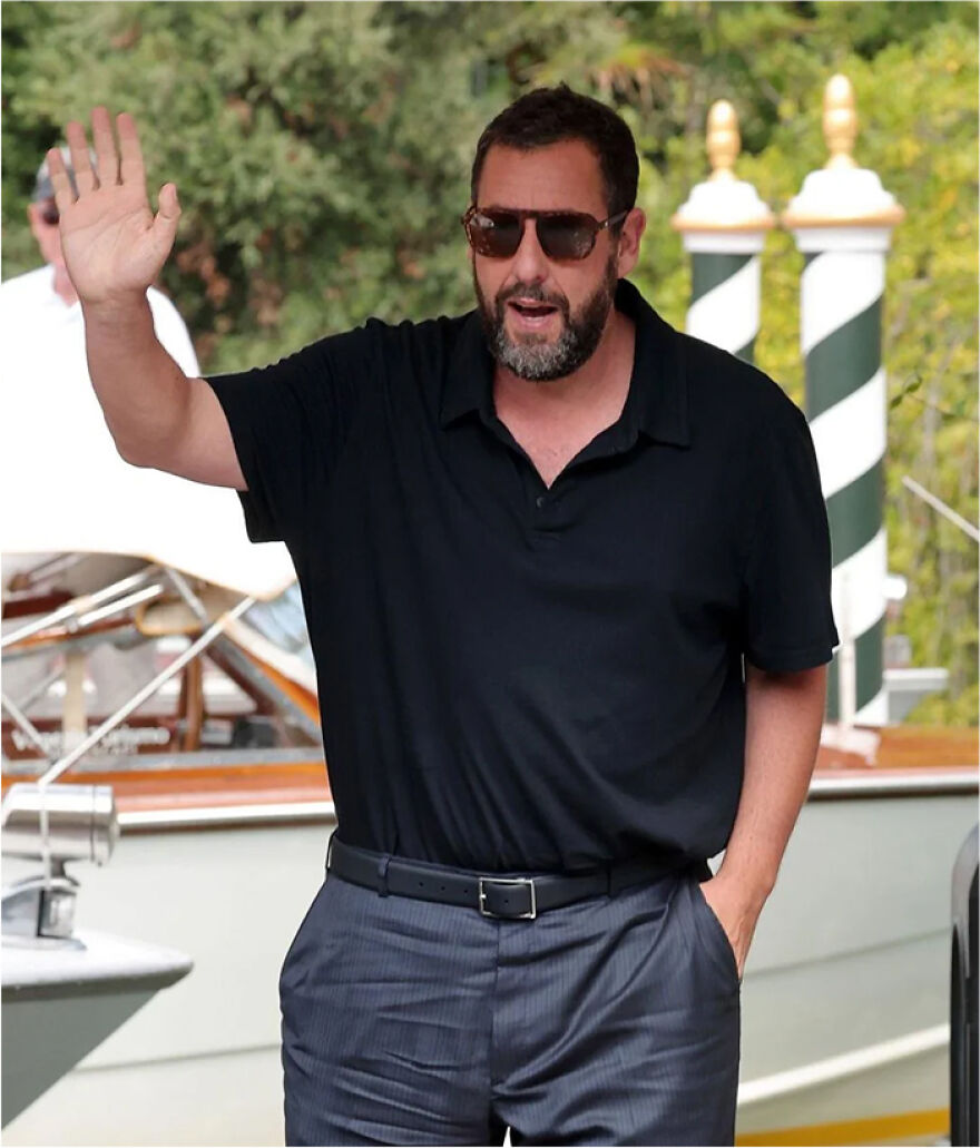 Adam Sandler wearing sunglasses, black polo shirt, and striped pants while waving outdoors by a dock.