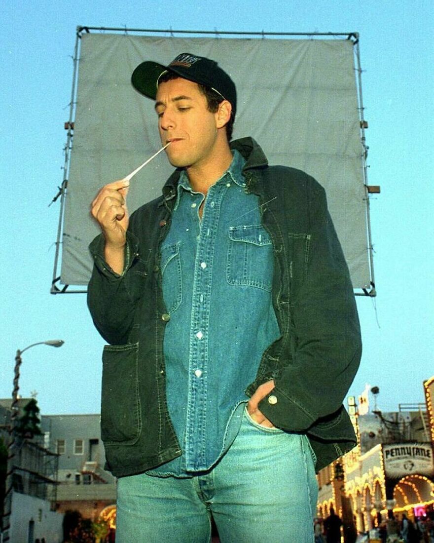 Adam Sandler wearing layered denim and a dark jacket, chewing gum with a casual street backdrop in daylight.