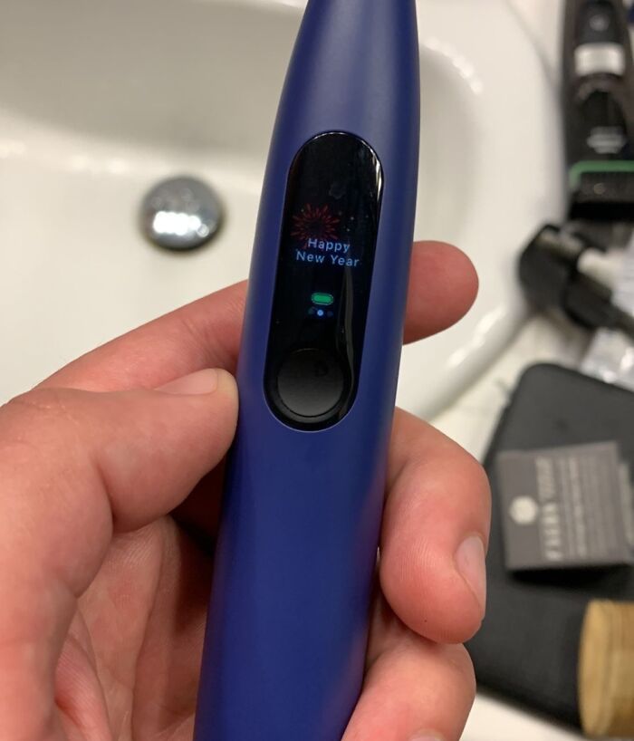 Hand holding a blue electric toothbrush displaying a Happy New Year message in a bathroom sink setting, dystopian scene.