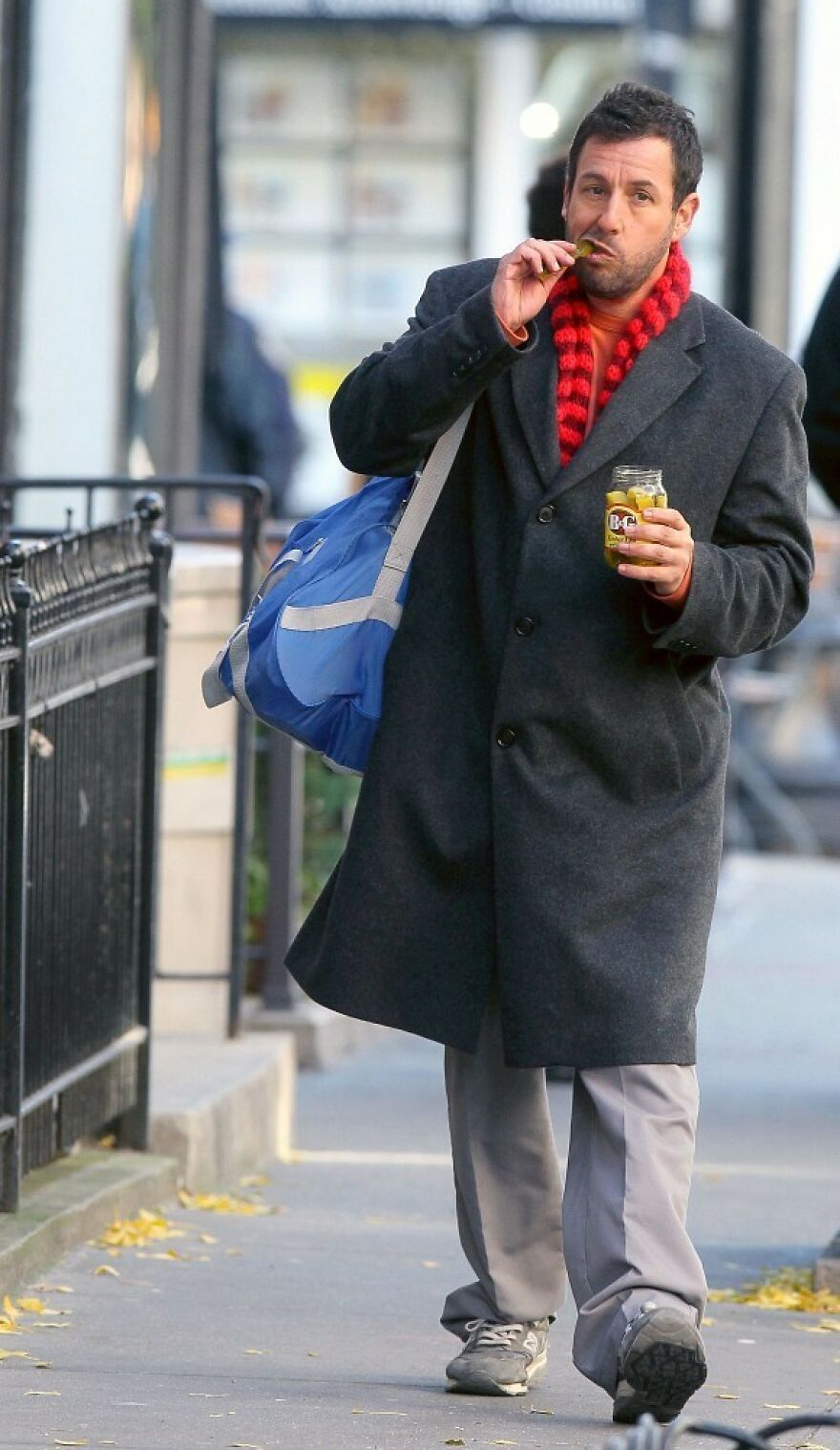 Adam Sandler wearing a dark overcoat and red scarf, carrying a blue bag while eating pickles on a city sidewalk.