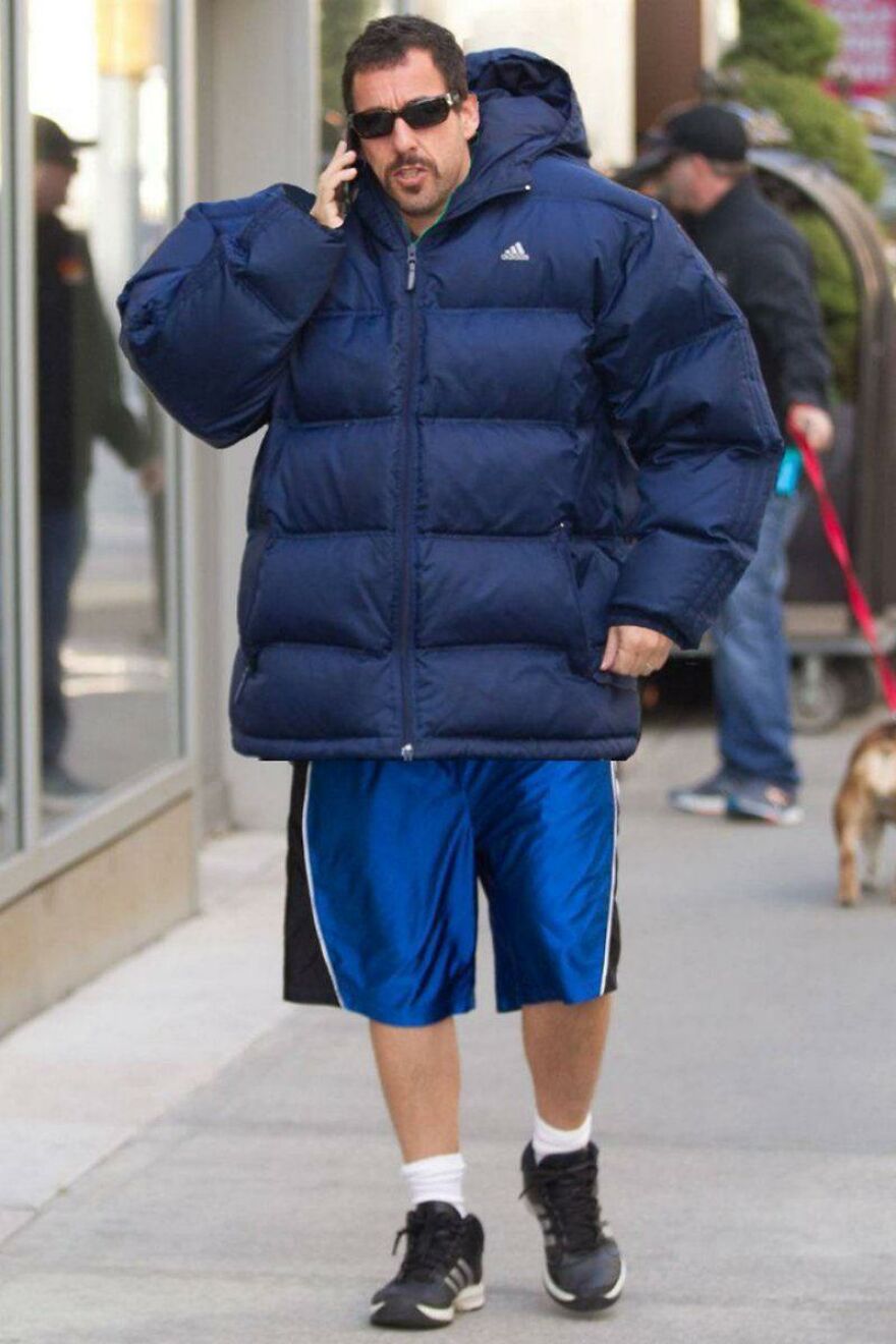 Adam Sandler wearing a large navy puffer jacket and blue shorts while walking and talking on the phone outdoors