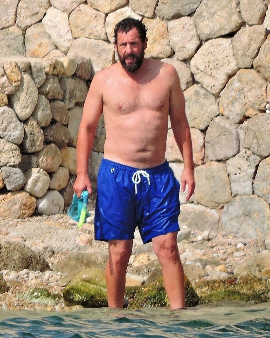 Adam Sandler wearing blue swim shorts standing in shallow water near a rocky shore with snorkeling gear.