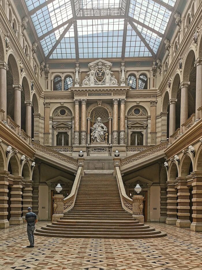 Grand interior architecture with a sweeping double staircase leading to a statue, surrounded by columns and ornate details under a glass ceiling.