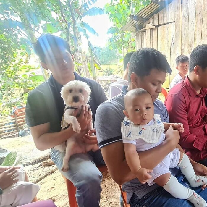 Hombre sosteniendo un perro pequeño y otro hombre cargando un bebé, momentos divertidos de perros capturados en cámara.