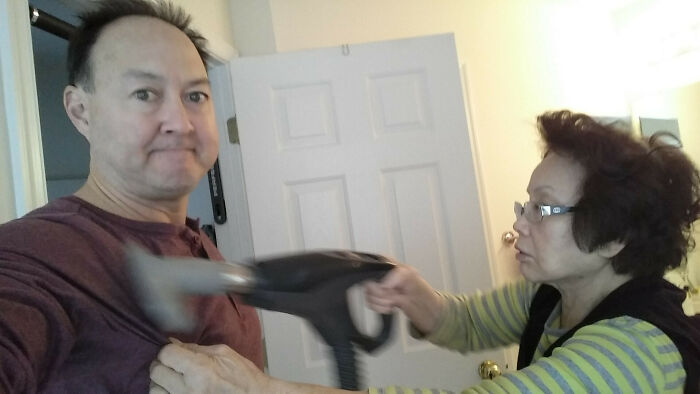 Middle-aged woman humorously steaming a man's shirt indoors, capturing a funny and wholesome parent moment shared by kids.