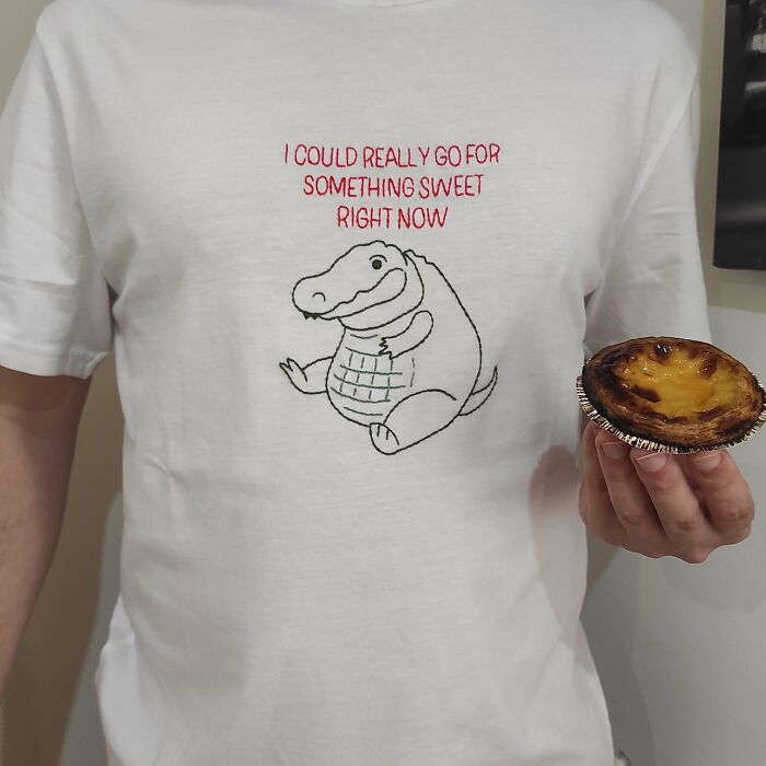 A person wearing a white t-shirt with a cartoon alligator and text, holding a pastry, featuring an embroidery creation.