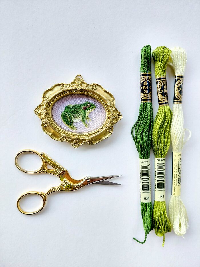 A stunning embroidery creation of a frog in a gold frame, with green embroidery floss and scissors, inspiring embroidery creations.