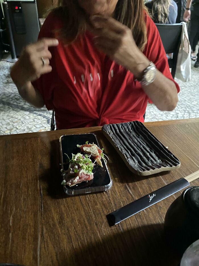 Mom in a red shirt sitting at a wooden table with food on a phone and a textured black plate nearby, capturing wholesome moments.