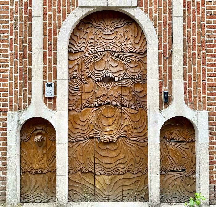 An elaborate wooden door with a wavy, textured design, flanked by two smaller arched doors, all set within a brick facade.