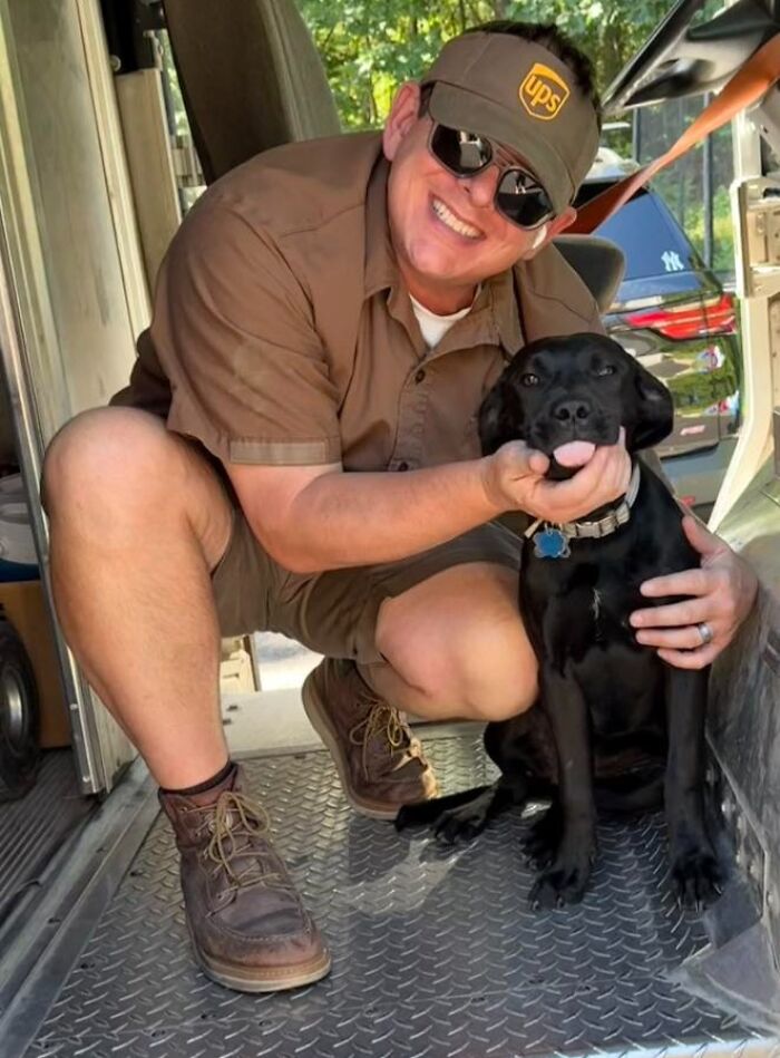 UPS driver smiling and crouching inside delivery truck while holding and posing with an adorable black dog.
