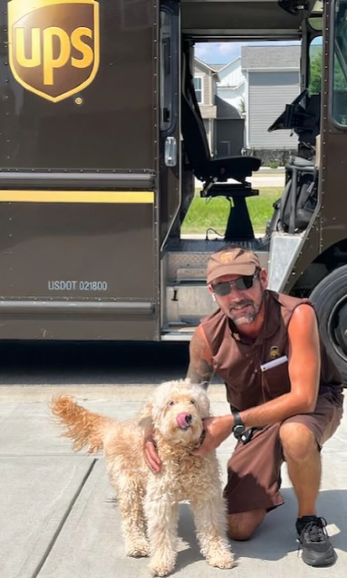 UPS driver kneeling beside an adorable dog outside a UPS delivery truck on a sunny suburban street.