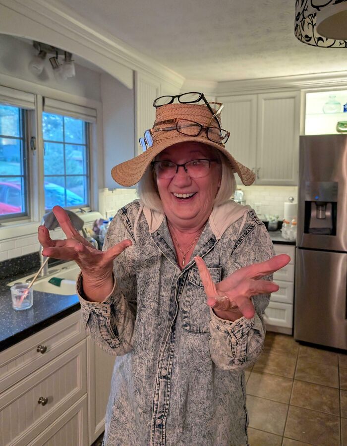 Funny parent wearing multiple pairs of glasses on a hat, smiling and gesturing in a bright kitchen setting.