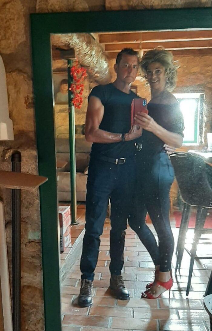 Couple dressed in black posing for a mirror selfie in a cozy rustic room, showcasing funny parents vibe.