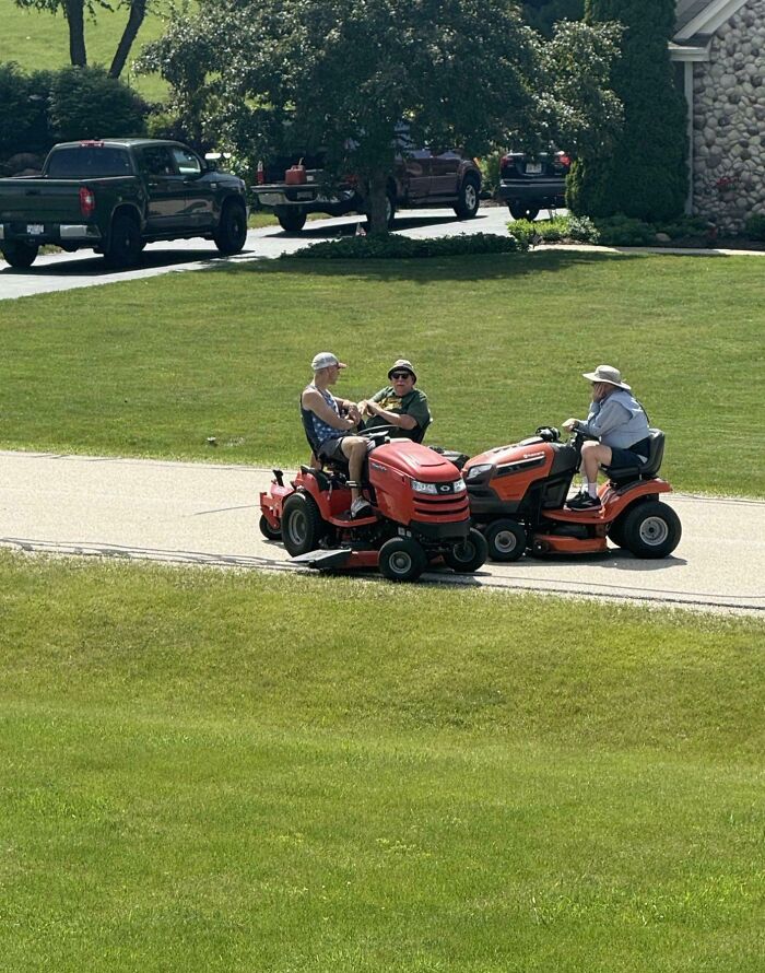 Three people riding lawn mowers facing each other on a driveway, showcasing funny parents enjoying a playful moment outdoors.