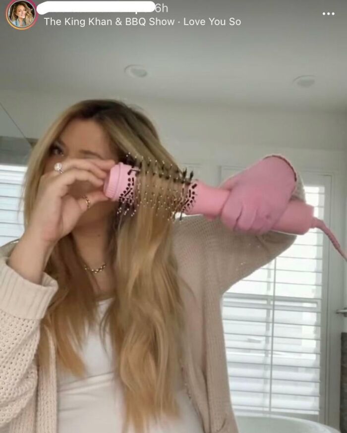 Woman influencer using a pink hair styling brush on long blonde hair, demonstrating beauty tips in a bright room.