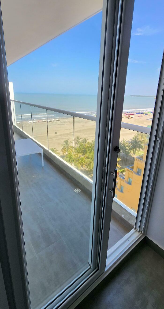 View from a vacation balcony with a locked sliding door overlooking an empty beach and ocean on a sunny day.