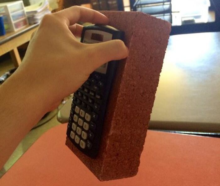 Hand holding a calculator attached to a brick as a humorous teacher's creative teaching tool in a classroom setting.