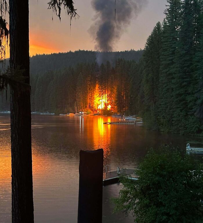 Forest fire near a lakeside cabin at sunset, illustrating vacation worst luck with flames and smoke rising among trees.