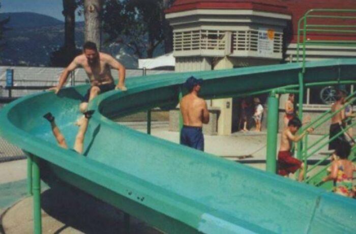 Two men and children playing on a large green slide at a water park illustrating funny differences between moms and dads.