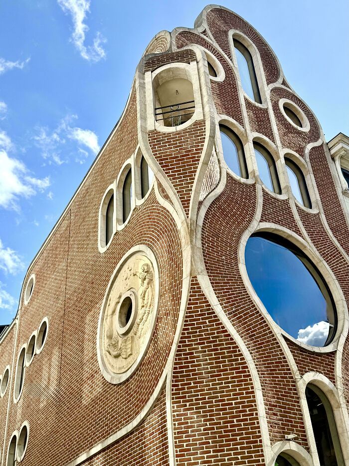 Unique architecture with undulating brickwork and organic shapes, featuring circular and arched windows against a bright blue sky.