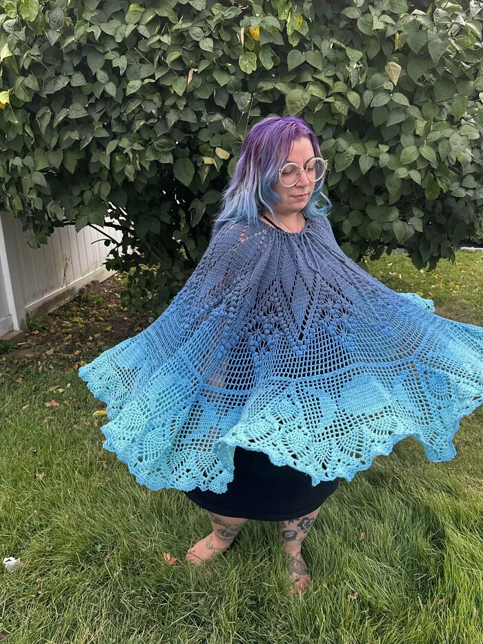 Person with purple and blue hair wearing a detailed blue crocheted shawl standing outdoors on grass near a leafy tree.
