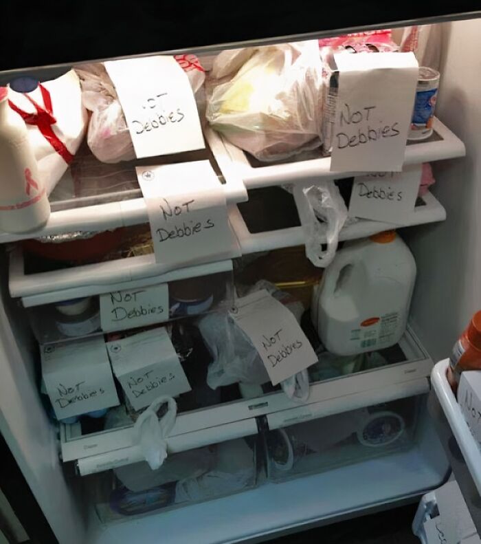 Office fridge cluttered with passive-aggressive notes labeled not Debbies highlighting coworker tensions.