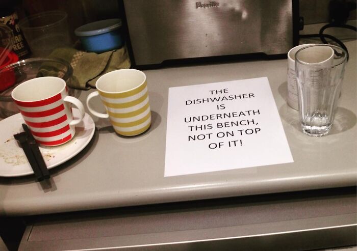 Passive-aggressive coworker note about dishwasher placement on a kitchen counter with cups and glasses nearby