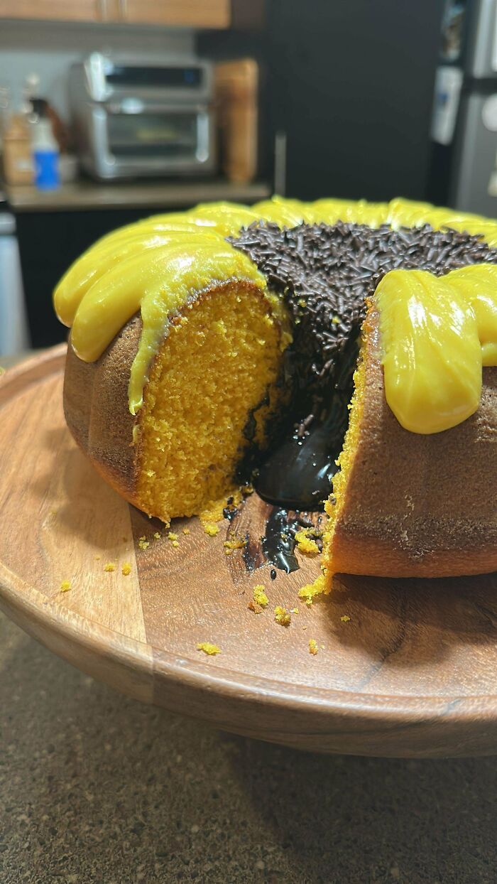 Yellow bundt cake with chocolate filling oozing out, topped with glossy yellow frosting and chocolate sprinkles on wooden plate.