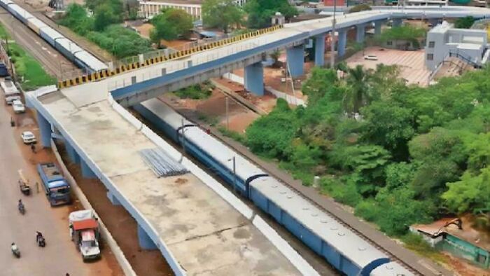 Aerial view of a blue passenger train traveling under a curved concrete bridge surrounded by trees and buildings.
