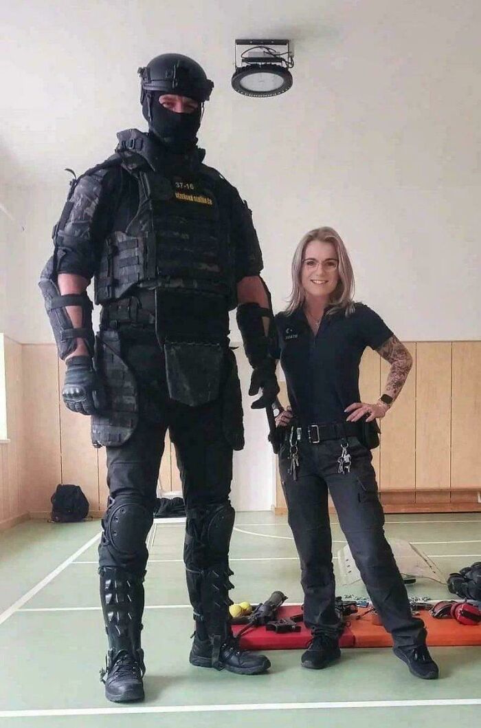 A very tall officer in tactical gear stands next to a smiling woman in black uniform inside a gymnasium setting.