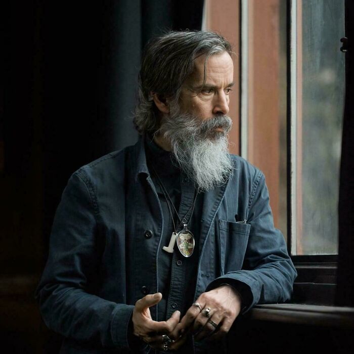 Bearded man with long gray hair gazing thoughtfully out a window, capturing a quiet moment at sunrise.