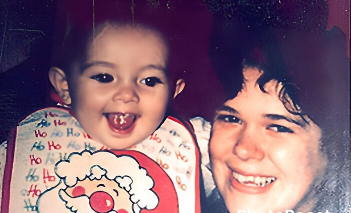 Baby wearing a festive bib with Santa design smiling joyfully next to a smiling adult, capturing surprising undiscovered moments.