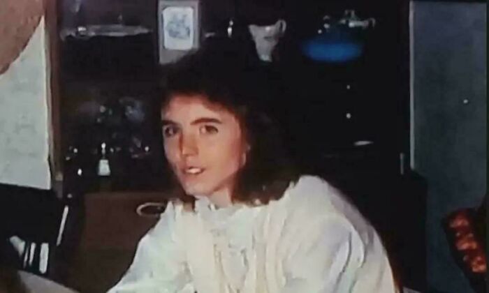 Young woman with long brown hair wearing a white blouse in a dimly lit room with shelves behind her, still remains undiscovered.